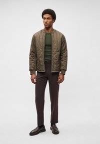Casaco bomber verde-oliva acolchoado com fecho de correr frontal, usado sobre uma camisa verde-escura justa e calças castanhas, combinado com mocassins escuros.