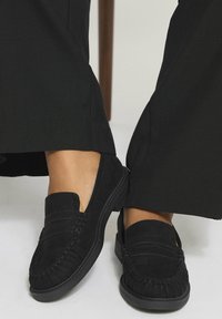 Mocassini in camoscio nero con punta arrotondata, cuciture dettagliate e suola in gomma, abbinati a pantaloni neri a gamba larga.