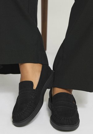 Mocasines de ante negros con punta redonda, costuras detalladas y suela de goma, combinados con pantalones de pierna ancha negros.