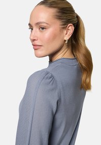 Femme aux cheveux blonds attachés en queue de cheval, portant des boucles d'oreilles créoles dorées et un haut en tricot bleu clair avec des manches bouffantes texturées, regardant sur le côté.