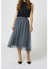 Femme portant un haut noir sans manches et une jupe gris fluide longueur genou, avec des escarpins noirs pointus, debout sur un fond blanc.