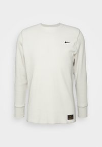 Vit långärmad tröja i våffelstickning med rund halsringning, Nike-swooshlogga på bröstet och svart och orange märktag nära fållen.