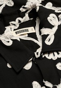 Tissu noir avec broderie florale texturée blanche et col, avec une étiquette beige portant l'inscription "WOODBIRD" en lettres grasses.