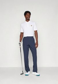 Biała koszulka polo z małym żółtym logo, zestawiona z granatowymi spodniami do golfa. Strój uzupełniają białe buty z niebieskimi akcentami oraz kij golfowy.