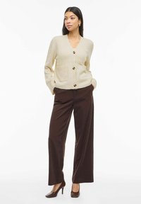Žena u bež kardiganu s dugmadi, tamnosmeđu širokih hlača i smeškim visokim potpeticama, stoji s rukama u džepovima na bijeloj pozadini.