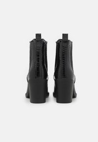 Une paire de bottines noires en cuir, avec un motif crocodile et des talons blocs, vues de l'arrière sur un fond blanc uni.