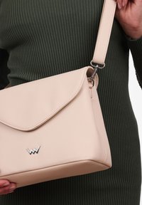 Beige křížová taška vyrobená z textura materiálu s překlopeným víkem a stříbrným logo akcentem, s nastavitelným popruhem a čistými liniemi.