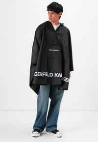 Een jongeman in een zwart Karl Lagerfeld regenponcho, blauwe jeans en zwart-witte sneakers, staand tegen een witte achtergrond.