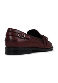 Mocassins de couro castanho-escuro com uma sola preta baixa, apresentando uma fivela decorativa e detalhes de costura ao longo dos lados. Textura suave em toda a peça.