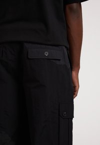 Schwarze Cargo-Hosen aus strukturiertem Stoff. Verfügen über eine Seitentasche mit einem Knopf und einem klaren, schlichten Design. Keine sichtbaren Muster oder Akzente.