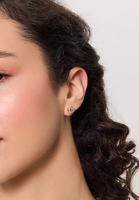 Złote kolczyki w kształcie obręczy mają splątany wzór z małymi, przezroczystymi kamieniami. Zbliżenie ukazuje szczegóły tekstury i krzywizn.