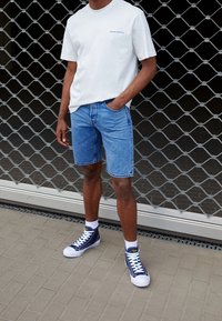 Camiseta de algodón blanca con un estampado azul, pantalones cortos de mezclilla azules y zapatillas altas marinas con suelas blancas sobre un pavimento gris texturizado.