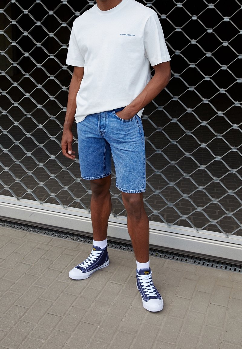 Camiseta de algodón blanca con un estampado azul, pantalones cortos de mezclilla azules y zapatillas altas marinas con suelas blancas sobre un pavimento gris texturizado.