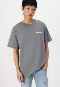 Szara koszulka z krótkim rękawem i okrągłym dekoltem, z małym jasnooliwkowym logo "Wrangler" na piersi. Noszona z wyblakłymi niebieskimi dżinsami.