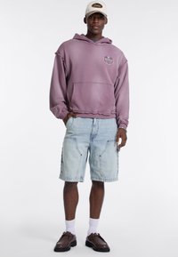 Felpa oversize viola con tasca frontale, abbinata a pantaloni cargo in denim chiaro. Scarpe marroni con lacci e calzini bianchi a metà caviglia completano il look.