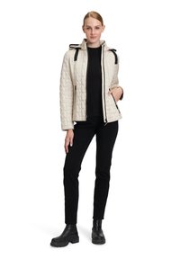 Giacca trapuntata color crema con dettagli neri e zip; presenta un cappuccio e tasche. Abbinata a un top nero e pantaloni neri, e stivaletti neri alla caviglia.