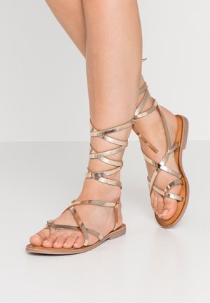 Füße, die metallisch roségoldene Gladiator-Sandalen mit dünnen, sich kreuzenden Riemen tragen, die um die Knöchel gewickelt sind, und flachen braunen Sohlen.