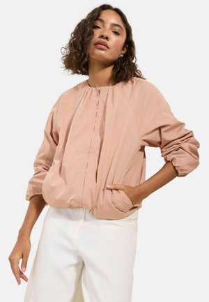 Frau mit mittellangem lockigem Haar, die eine hellrosa Reißverschlussjacke mit gerafften Ärmeln und weiße Hose trägt, Hand in der Tasche, vor weißem Hintergrund.