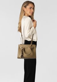 Handtasche aus cognacfarbenem Leder mit strukturierter Form, geflochtenem Griff, Metallbeschlägen und abnehmbarem Anhänger. Verstellbarer Schulterriemen inklusive.