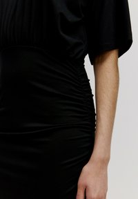 Schwarzes Kleid mit kurzen Ärmeln, gerafften seitlichen Details und figurschmeichelndem Schnitt. Der Stoff wirkt glatt und elastisch.