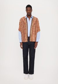 Camisa de manga corta con estampado en naranja, con acentos en amarillo y blanco, superpuesta sobre una camisa de botones azul clara. Combinada con unos jeans oscuros y zapatillas blancas.