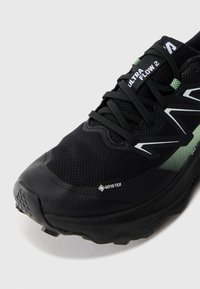 Black athletic shoe with mesh upper, GORE-TEX label, green accents, and textured grip sole. Features reinforced toe and structured lacing.