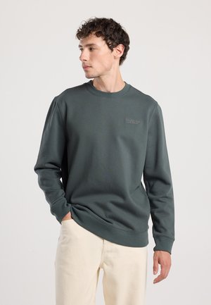Sudadera verde oscuro, de cuello redondo, con puños y dobladillo acanalados, que presenta el texto "PARQUE NACIONAL" en un tono más claro en el pecho. Combinada con pantalones claros.