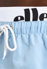 Lys blå badebukser med en hvid elastisk talje, der har sort tekst, justerbar snøre og en glat tekstiloverflade.