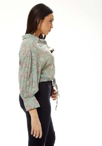 Green floral blouse with puff sleeves, cinched at the waist with a tie. Features a buttoned collar and intricate pink and white patterns.