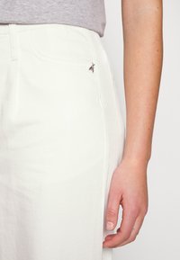 Jeans blancos con un bolsillo delantero que presenta un pequeño detalle de abeja plateada, combinados con una camisa gris; textura suave y ajuste entallado.