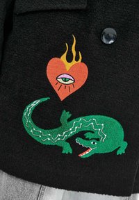 Zelený vyšívaný krokodíl a horiace srdce s okom nad ním na čiernej textúrovanej látke blízko gombíka.