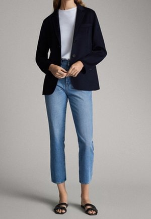 Woman wearing a black blazer, white top, light blue straight-leg jeans, and black open-toe sandals, standing against a plain background.