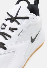 Hvide atletik sko med tekstureret mesh og syntetisk læder. Har sorte accenter, sorte snørebånd og et "Just Do It"-mærkat i sort på siden.