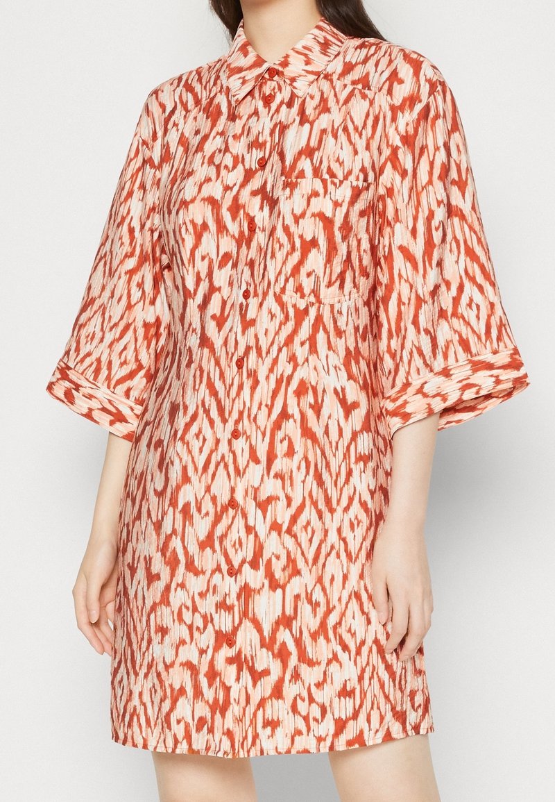 Femme portant une robe chemise boutonnée mi-longue aux manches 3/4, en tissu à motif abstrait orange et blanc.