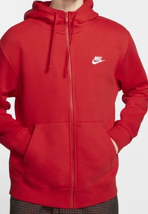 Person wearing a red Nike zip-up hoodie with a hood and front pockets, hands partially inside pockets, against a plain background.