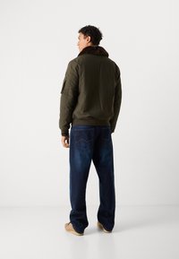 Olivenfarvet bomberjakke med en brun pelskrave, parret med løstsiddende mørkeblå jeans og beige fodtøj. Har skulderlomme.