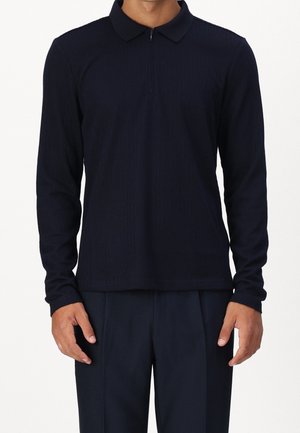 Male model wearing a long-sleeve navy blue polo shirt with a front zipper and matching navy pants, standing against a white background.