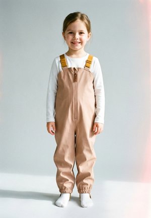 Lächendes junges Mädchen, das beige Latzhose mit gelben Trägern und ein weißes Langarmshirt trägt, barfuß vor einem schlichten Hintergrund steht.