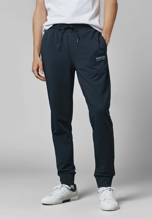 Mand iført sorte Red Bull Racing joggingbukser med snørelukning i taljen og hvide sneakers, stående med den ene hånd i lommen mod grå baggrund.