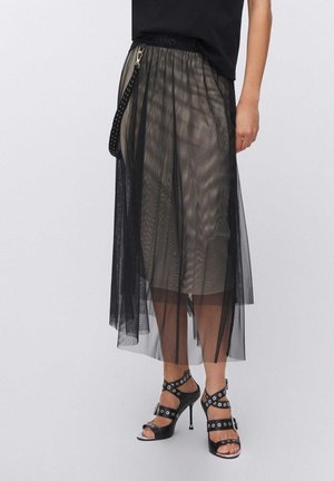 Donna con una gonna midi a strati in tulle nero con una fodera beige, abbinata a sandali neri con tacco alto e borchie, e una maglietta nera.