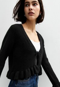 Schwarze, flauschige Strickjacke mit V-Ausschnitt, Bindeband und Rüschenabschluss. Lange Ärmel reichen bis zum Handgelenk und sind über einem weißen Oberteil geschichtet.