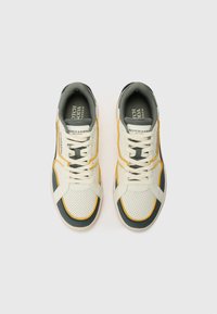 Sneakers met een crèmekleurige bovenkant met geperforeerde structuur, grijze accenten en gele stikdetails, die een modern sportief ontwerp tonen.
