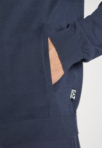 Sweater azul-marinho feito de tecido texturizado, com um bolso lateral e punhos canelados. Um pequeno logótipo EA7 é visível na parte inferior.