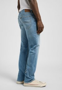Los jeans de mezclilla en un tono azul claro presentan un diseño de pierna recta, detalles de bolsillos tipo parche y una etiqueta de cuero marrón en la cintura.