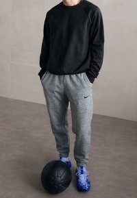 Personne portant un sweat-shirt noir, un pantalon de survêtement gris et des chaussures de sport bleues, debout à l'intérieur les mains dans les poches, avec un ballon médicinal noir près des pieds.