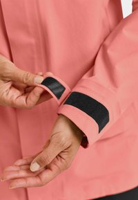 Pink waterproof jacket with a black Velcro cuff. A hand adjusts the Velcro strap at the sleeve, revealing smooth fabric texture.