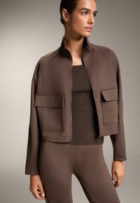 Rjava kratka jakna iz gladkega blaga, ki ima dva sprednja žepa, visok ovratnik in zadrgo, v kombinaciji s temnim rebrastim puloverjem.