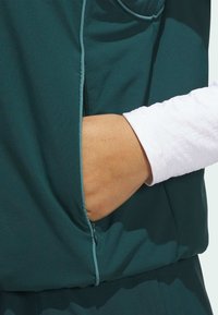 Sinivihreä urheilutakki vetoketjullisella taskulla ja alla teksturoitu valkoinen pitkähiha-paita. Sileä kangas, jossa kontrastitikkaukset.