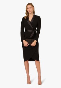 Schwarzes Wickelkleid mit langen Ärmeln, satinierter Kragen und Gürtel. Glatte Textur, knielang mit einem seitlichen Schlitz, kombiniert mit einer plissierten Clutch.