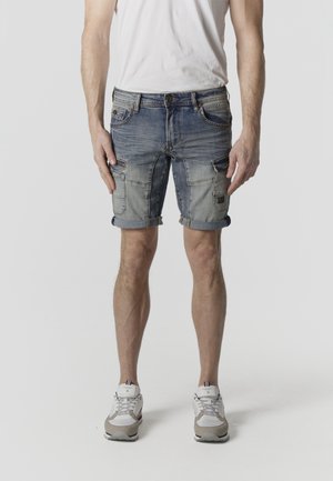 Hombre vistiendo camiseta blanca, pantalones cortos cargo de mezclilla azul remangados y zapatillas blancas, de pie sobre un fondo claro y liso.